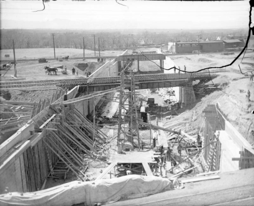 View of Alameda underpass construction in Denver, Colorado; shows concrete forms, railroad bridges, machinery, sheds, and men working.