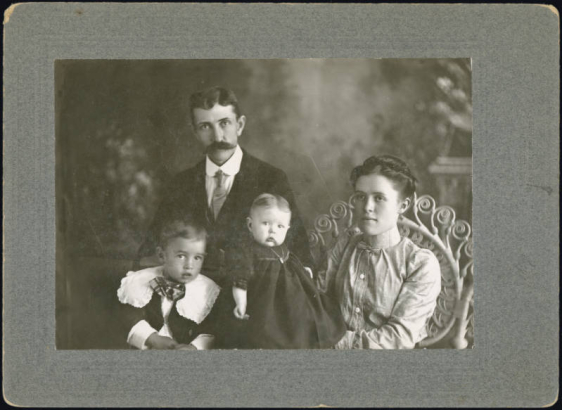 Studio portrait of Pablo C. de Baca, his wife Emilia Rudolph C. de Baca, their unidentified son and an unidentified infant. Pablo wears a dark suit and tie, has his hair parted in the middle and has a waxed mustache. His wife Emilia wears a blouse with puffed sleeves. The young boy wears a dark vest and a shirt with a lace Bertha collar and an over sized bow tie. The baby wears a dark dress and has its hair parted in the middle.