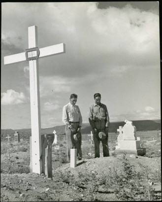 Two men stand at the grave of Maria Ursulita Vialpondo in the cemetery in Chama (Costilla County), Colorado. An elderly Hispanic American man stands with his hat in his hand. Beside him stands possibly his son, Moises Vialpondo. A large cross is near the men. The gravestone had a lamb on top and a heart shaped center with letters that read: "Maria Ursulita Vialpondo July 29, 1877, Oct. 21, 1940."