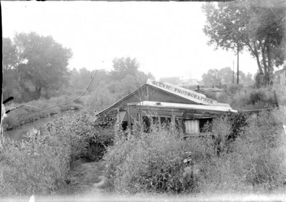 View of the home and photography studio of Charles S. Lillybridge, in Denver, Colorado; shows the Archer Canal and a frame house with porch and sign: "Scenic Photographer." Industrial buildings show past trees.