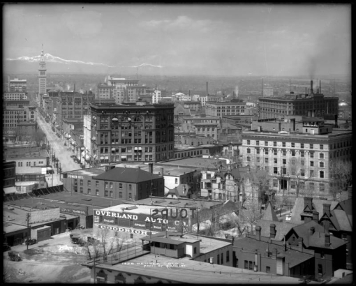 Right center of panorama view taken from Colorado State Capitol Building, Denver, Colorado; includes downtown business district with. Signs read: "Plymouth Hotel," "Tours," " Overland Auto Co.," "The Byron Motor Car Co.," "F. C. Ayres, Fireproof Grain Elevator," The Front Range with snow covered Longs Peak is in the distance.