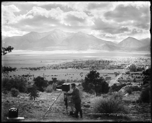 Evening view of Mount Princeton and Buena Vista (Chaffee County), Colorado; thought to be L.C. McClure standing behind camera & tripod; box of camera supplies, foreground left; Collegiate Range, Rocky Mountains, and dramatic clouds.