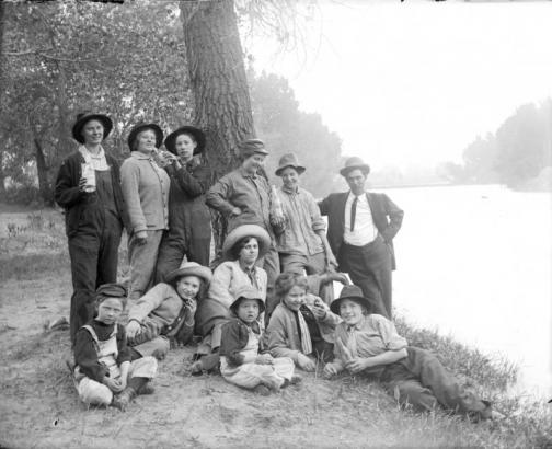 Outdoor portrait of a group of women dressed as men and a man near the South Platte River in Denver, Colorado. A few of the women hold bottles or corncob pipes.