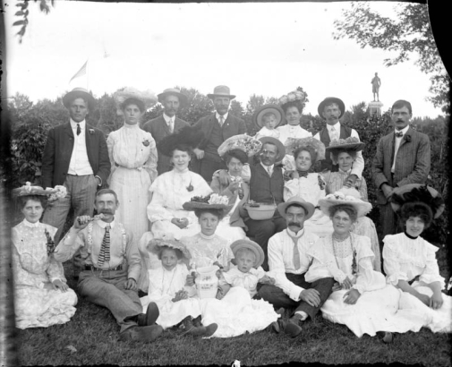 Outdoor portrait of a group of men, women, and children in City Park, Denver, Colorado. One woman holds a pitcher while another eats from a bowl. A statue of Robert Burns is in the distance.