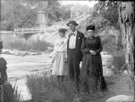 Outdoor portrait of a man and two women near a creek in the mountains of Colorado. The women wear hats decorated with flowers. A water tank and a wooden bridge are in the distance.