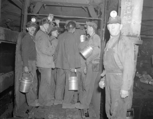 Miners stand in and near the cage of a coal mine in Colorado. They hold lunch pails and wear miner's lights.