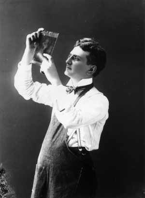 Self portrait of George Lytle Beam examining a glass plate. He wears a lab apron, striped bow tie, white shirt with wing-tip collar and his pince-nez eyeglasses.