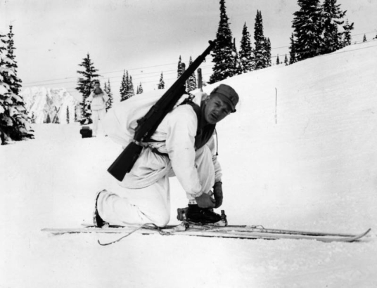 Tenth Mountain Division Corporal, Richard Cochran bends over his skis as though adjusting his bindings. He is wearing winter camouflage of "whites" as well as a knapsack and his rifle. Almost obscured behind him is  another soldier in the same uniform standing and facing directly toward the camera. Both are on a snow covered mountain in an unnamed location.