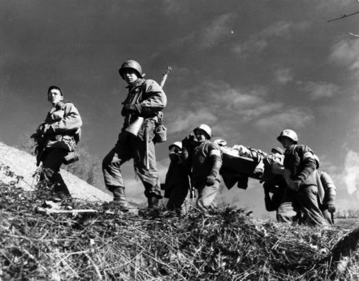 Two soldiers from the Tenth Mountain Division, 85th Regiment, 1st Battalion, walk in front of a wounded soldier on a litter carried by five others. According to the caption the soldier had been wounded in the attack on Mount Belvedere during the Italian Campaign.