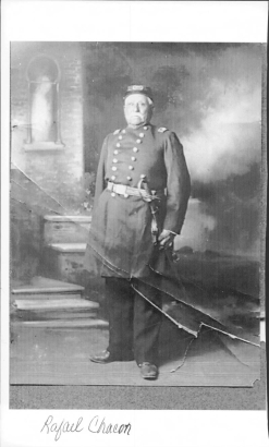 Studio portrait of Major Rafael Chacón (retired). He is dressed in his United States Army uniform. His jackets has two rows of brass buttons, he wears a cap and has a sword at his waist. He stands in front of a painted backdrop.