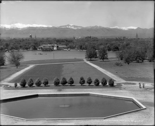 View of range of Rocky Mountains, including 14,260-foot Mount Evans at left background, from City Park in Denver, Colorado; shows octagonal fountain pool, landscaped lawn with pedestrians on pathway, lake with pavilion and bandshell, and the skyline of downtown Denver is visible.