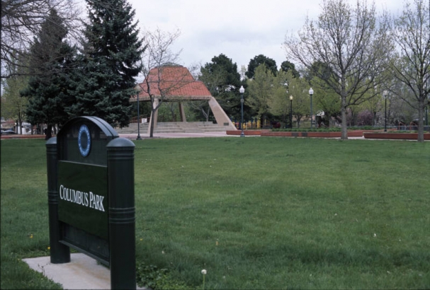 View of the sign for Columbus Park between West 38th (Thirty-eighth) and West 39th (Thirty-ninth) Avenues and Navajo and Osage Streets in the Sunnyside Neighborhood, Denver, Colorado. The sign reads: "Denver Department of Parks and Recreation Columbus Park." A picnic shelter, an Aztec inspired pavilion with a red stepped pyramid shaped roof and stucco supports, is in the distance.