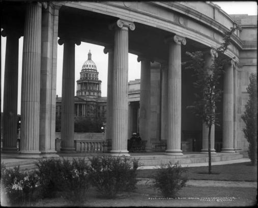 View of the Colorado State Capitol in Denver, Colorado; the capitol is framed by columns of the Greek Theater and Colonnade of Civic Benefactors in Civic Center Park (completed in 1919); two people sit on bench under colonnade.