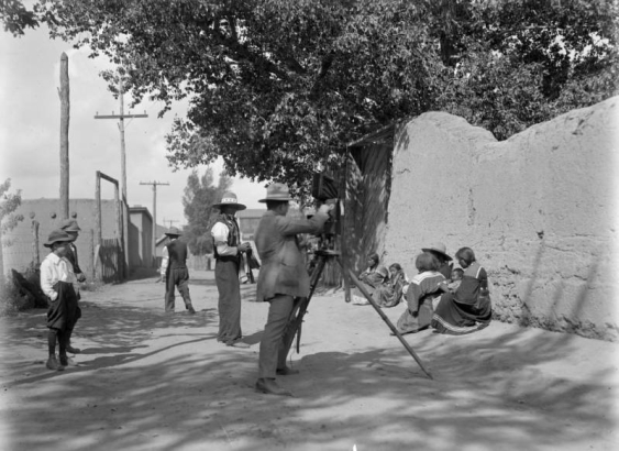 View of a photographer with tripod and camera, and his subjects, Native American men, women and children, in Taos, New Mexico.