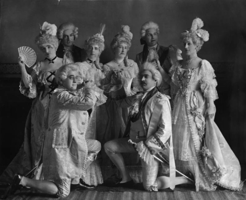 A group poses in 18th century ball costumes and powdered wigs, Colorado Springs, Colorado; women wear lace gowns, jewerly, plumes and hold fans, men have on jackets with tails, tights, knickers and ruffled shirts. Members include: Zeph Hill, (Mrs. Theo) F. P. Holland (?) Frank Wordmarch, Xianau Lee (married Tarbor Hagam,) Julian Streans (Mrs. T. B.,) Mrs. E. F. Powers and A. Vermis.