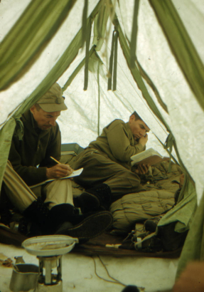 View of Captain Albert Jackman and Lieutenant Paul Townsend of the 87th Mountain Infantry Regiment (re-enforced) inside a green Everest Tent during the Mount Rainier Test Expedition. Jackman is taking notes and Townsend is reading. Equipment includes a stove, boots and crampons, and a sleeping bag. Jackman is wearing a cap, a dark shirt, khaki pants, and smooth-soled black boots. Townsend is wearing khaki shirt and pants and smooth-soled black boots.
