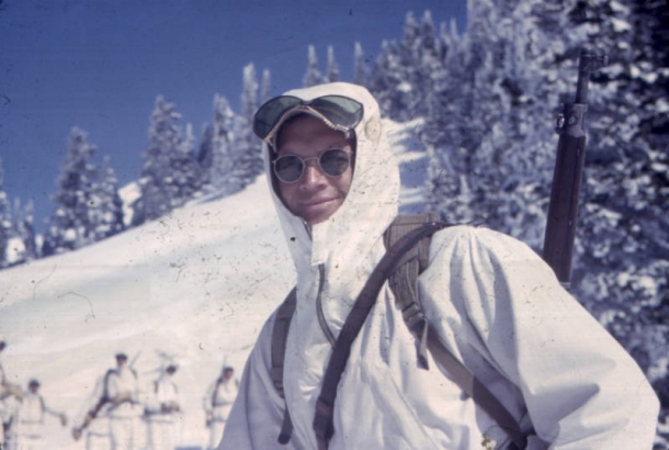 Portrait of Paul Lafferty, Captain in the 87th Mountain Infantry Regiment (re-enforced). He is wearing a white anorak, sun glasses, goggles,  a knapsack and has a rifle over his shoulder. In the background other skitroops dressed in the same uniforms stand at the base of a snow covered hill in the mountains near Paradise, Washington.