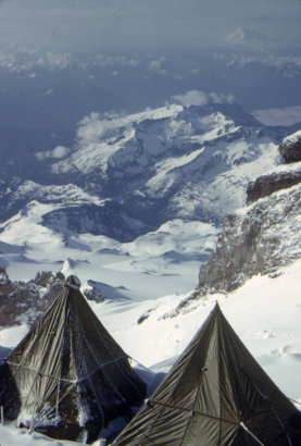 View of Camp Two looking through Cadaver Gap at Camp Muir, (also known as Camp One during the Mount Rainier Test Expedition conducted by men from the 87th Mountain Infantry Regiment, re-enforced). Two pyramid tents are pitched in the snow in the foreground. The stone cabin at Camp Muir is visible on a slight rise across a large open snow field. Beyond Camp Muir rough mountain territory stretches into the distance.