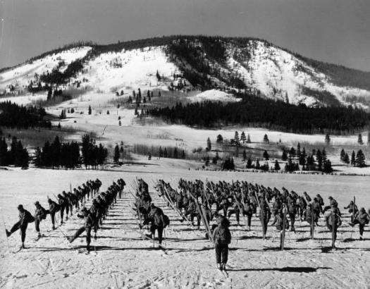 Still from the Warner Brothers film: "Mountain Fighters." Approximately one-hundred soldiers from the Tenth Mountain Division, 87th Mountain Infantry Regiment, Company L, execute an about face on skis under the command of Lieutenant William J. Bourke on a flat snow-covered field. In the background, smaller groups of skiers are visible training in the trees at the base of one of the mountains surrounding Camp Hale, Colorado.