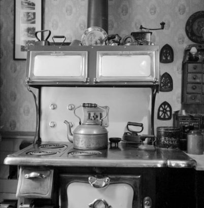 A stove and its stovepipe are in front of a wall with patterned wallpaper. The stove has six burners, and a kettle rests on one of them. The range has an oven with an enamel door and two irons sitting on it. Two warmers are aboe the range. Bric-a-brac such as a coffee grinder, irons, and plates, decorate the top of the stove. Pictures and other ornaments hang on the walls. A coffee tin is to the right of the stove.