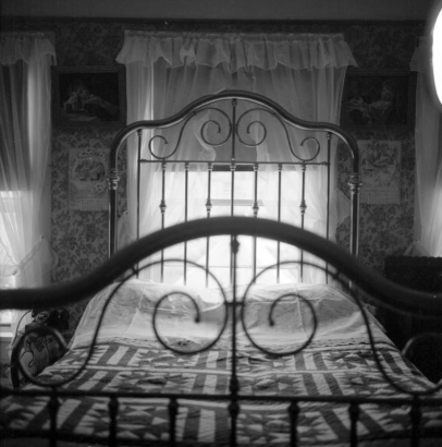 A  double bed in the Prosser house is illuminated by the light streaming in through the lace curtains of the window behind it. The brass headboard against the window is decorated with spindles and curved brass. The brass footboard is arched and has decorative brass pieces inside it. A quilted spread is on the bed.