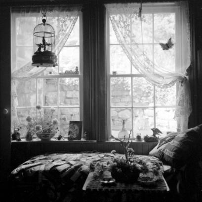 The sun streams through two windows of a Central City bedroom where a twin bed with a quilted cover is in the foreground. The windowsills next to the bed are decorated with bric-a-brac, including flowers, vases, bells, and miniature birds. A decorative birdcage with a bird in it hangs in front of the window. Lace curtains, gathered at the sides, decorate each window. A butterfly design is on the curtain on the right. The masonry wall and the vegetation in the yard are visible through the window.