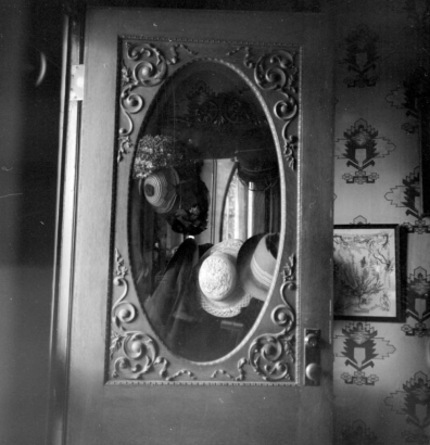 Straw and cloth hats hanging on a hatrack are reflected in an oval mirror in the Prosser house. Sunlight illuminates the left side of the image. The mirror has a rectangular frame and scroll-work around the edges. Another mirror is visible in the reflection of the mirror. An embroidered, framed picture hangs to the right. The wallpaper has a decorative pattern on it.
