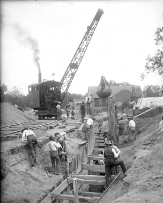 View of (probably) Lipan Street in Denver, Colorado; shows a ditch with shoring, and men working with a clam shovel crane.