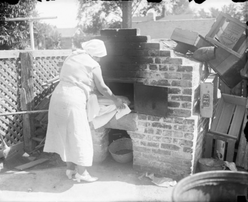 A woman bakes bread in an outdoor brick oven in Denver, Colorado. Wooden crates near the oven read: "Snowy Range Mountain Lettuce Burton Produce Co. Denver, Colo." and "AFG".
