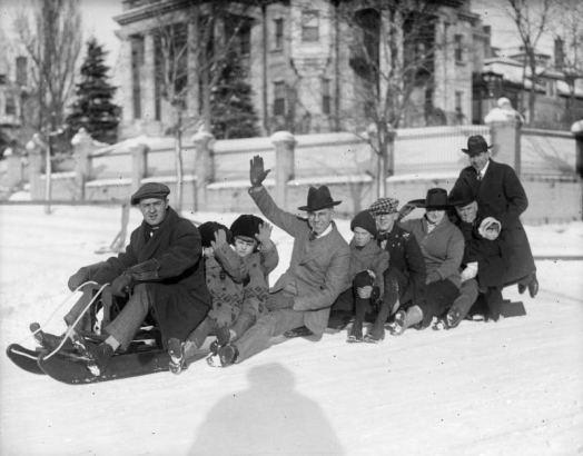 Men, children and a woman pose on a sled near the David Moffat mansion at 808 Grant Street in Denver, Colorado; shows Bert Rhoads (1st on sled), Charles Gates (waving), Hazel Rhoads Gates (holding onto her hat) and Harry Fisher Rhoads (holding one of the Gates' daughters).