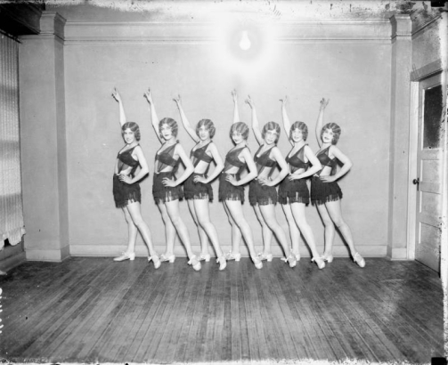 Dancers from the Georgia Land School of Dancing pose for a group photo in Denver, Colorado. The women, part of the Georgia Lane Group, wear short fringed skirts, bikini tops with sheer fabric overlays, and  have their hair marcelled (waved). The woman posed at the extreme left is Sarah Francis "Sally" Orr.