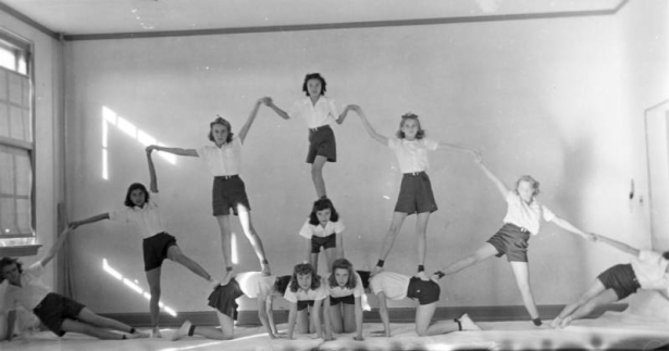 An eighth grade girl's gym class forms a human pyramid at the Central School in Alamosa, Colorado. The girls wear white, short sleeved blouses, belted dark shorts, socks and white tennis shoes. Four girls are on their hands and knees in the center. Another girl is on her hands and knees on the top center of the four girls. Another stands on her back, with others standing, holding hands with outstretched arms, to form a large arch that spreads across the room.