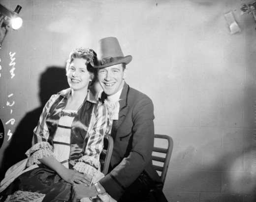 A man and a woman dressed in costume for Saint Patrick's Day pose, probably Denver, Colorado. She wears a dress with ruffled sleeves, lace and satin bodice, and a tiara. He wears a hat and suit with satin and wide lapels.