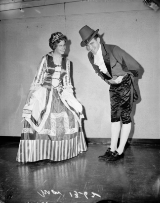 A man and a woman dressed in costume for Saint Patrick's day pose for a photo. She wears a long patterned dress with ruffled sleeves, lace and satin bodice, and a tiara. He wears a hat, suit with satin and wide lapels, knickers and tights.