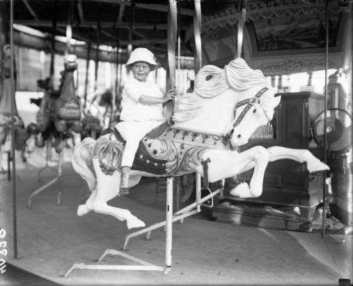 A young girl rides a carousel horse on the merry-go-round at the Lakeside Amusement Park in Denver, Colorado. The child wears a white pants and blouse, button shoes, and a hat.