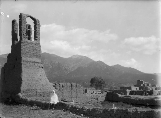 View of mission ruins and graves at Taos, New Mexico; a Native American (Taos Pueblo) poses in a blanket. Adobe pueblo dwellings and Pueblo Peak are in the background.