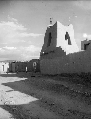 View of Taos Pueblo, New Mexico; shows Native American (Taos Pueblo) adobe walls, a tower with arches, and a lamp.
