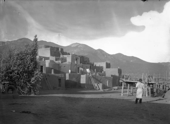 View of Taos Pueblo, New Mexico; shows a Native American (Taos Pueblo) man, scaffolds, adobe pueblo dwellings, and drying laundry.