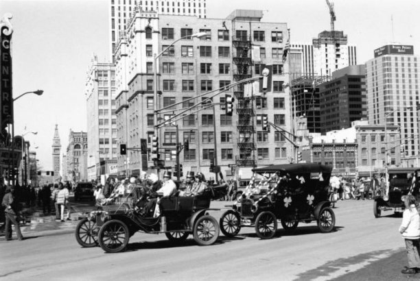 View of the Saint Patrick's Day Parade at 16th (Sixteenth) Street and Broadway in the Central Business District, Denver, Colorado. Men and a woman in hats ride in decorated antique convertible cars. The Paramount Theater and Daniels and Fisher Tower are in the distance.