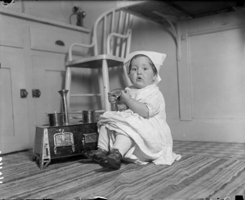 Harriet Rhoads, daughter of Harry M. Rhoads plays in the kitchen with her toy pots and stove, Denver, Colorado. Shows: cabinets, chair, rug.