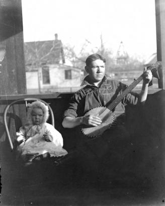 Portrait of a young man playing a banjo in Denver, Colorado, by a baby in a fur hood and lace dress.