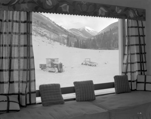 View (from inside) of snow, a mountain, and a tracklayer vehicle, probably in Colorado. Interior furnishings include a ledge seat, pillows, and a flower patterned valance.