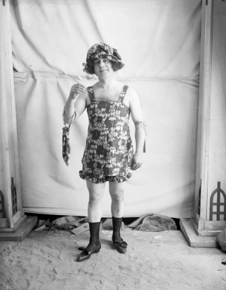 Photographer Harry M. Rhoads poses in costume. He wears a floral patterned women's bathing suit with a matching bonnet and hand bag, women's shoes and black socks.