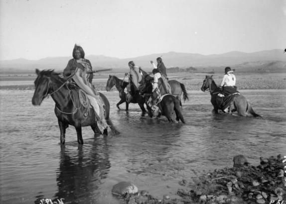 View of a Native American (Ute) scouting party mounted on horseback as they cross the Los Pinos river, La Plata County, Colorado; three men have rifles, one a pistol; men wear moccasins, fringed leggings, blankets, shirts, and braided hair; some have feathers in their hair; all horses have bridles and saddles.
