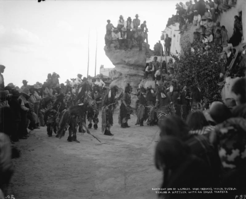 Native American men (Hopi), snake dancers, stand in a circle at Walpi Pueblo, First Mesa, Arizona; five men handle snakes, one holds a snake in his mouth; one man strokes a rattle snake (on the ground) with a feather, while groups of unidentified Native American and white men and women look on; snake members wear kilts and bead necklaces; some wear body paint.