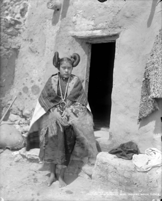 Outdoor portrait (standing) of a Native American (Hopi) woman near a Pueblo doorway at Walpi Pueblo, First Mesa, Arizona; she wears a dark manta, print and fringed shawl, woven blanket shawl, bead necklace with open hoop pendant, and hair in side whirls.