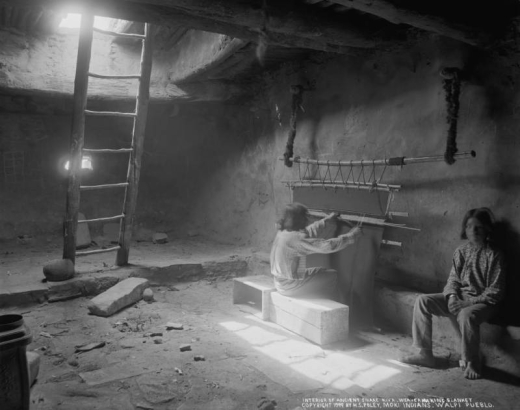 Interior view of a kiva, possibly the snake kiva, at Walpi Pueblo, First Mesa, Arizona; two unidentified Native American (Hopi) men sit near a loom; a wooden ladder leans against a large opening in the ceiling; a clay bowl is next to the ladder; one man sits on a wooden crate at a wooden loom suspended from the ceiling by fur pelts, and weaves a dark blanket or rug; one man sits against the wall; both men wear pants and print shirts; one wears no shoes.