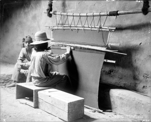 Interior view of a kiva, possibly the snake kiva, at Walpi Pueblo, First Mesa,  Arizona; two unidentified Native American (Hopi) men sit in the kiva; one man sits on a wooden crate at a wooden loom and weaves a dark blanket or rug while another looks on; both men wear pants and print shirts, one wears a woven hat, one wears a headband and no shoes.