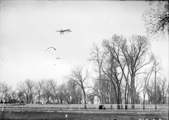 View of a Bleriot airplane in flight over Denver, Colorado, shows trees and fences.