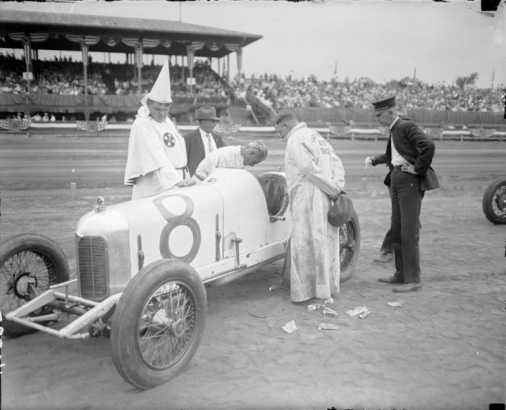 Ku Klux Klan members inspect the Miller Special race car owned by Ralph de Palma at Overland Park race track in Denver, Colorado. People crowd the bleachers. The driver, identified as Mr. Miller, wears a duster and holds a cap.